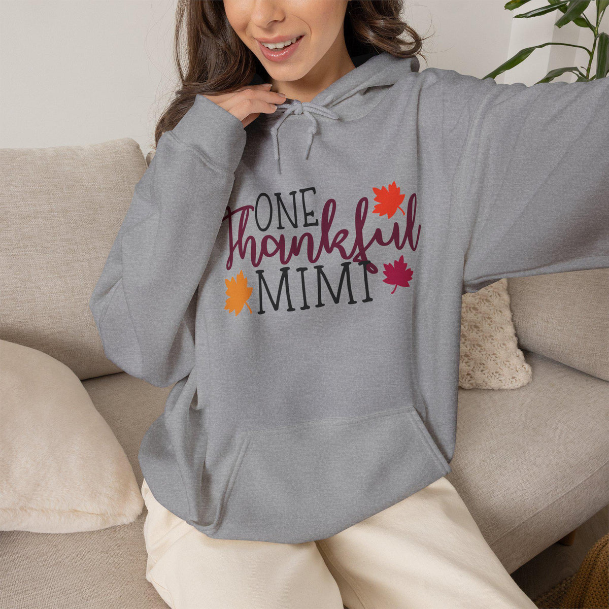 Woman wearing a gray hoodie featuring One Thankful Mimi lettering with colorful autumn leaves in a cozy living room seasonal women's fashion apparel