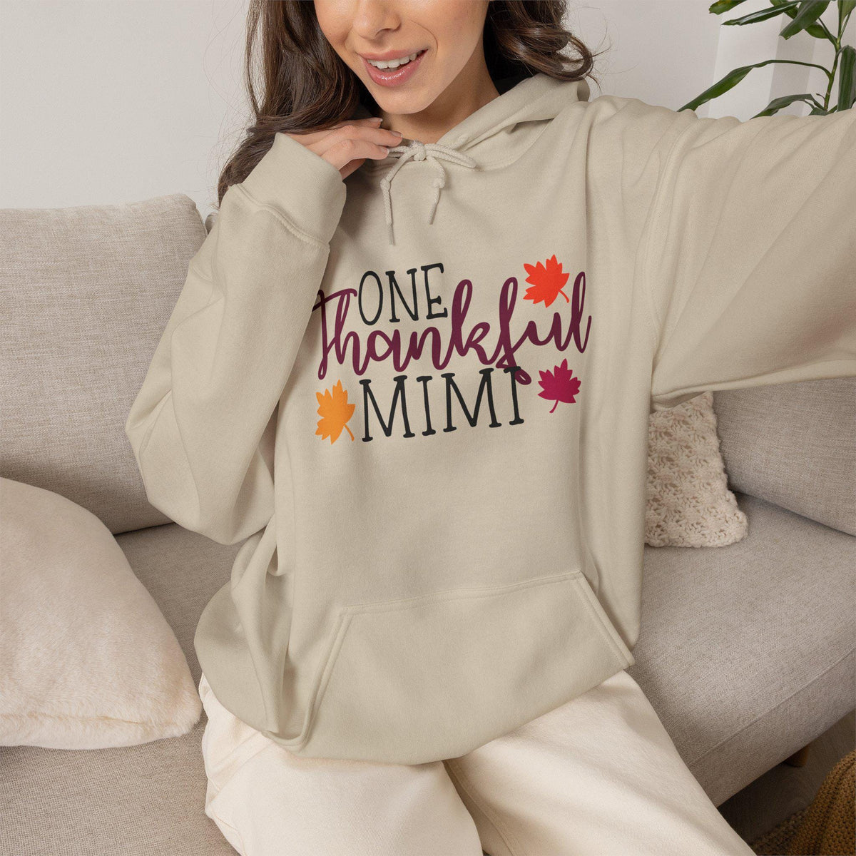 Woman modeling a cozy cream hoodie with One Thankful Mimi lettering and colorful autumn leaves for a warm seasonal fashion look