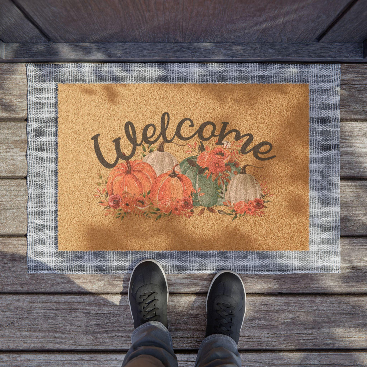 Rustic coir doormat with hand lettered Welcome text surrounded by pumpkins and autumn flowers framed by a gray plaid border on a wooden porch