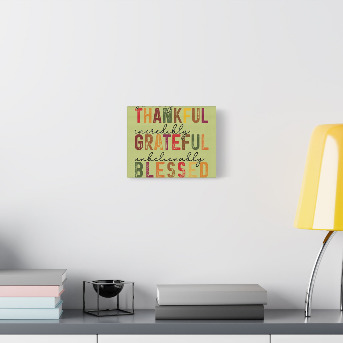 Distressed autumn wall canvas reading Thankful Grateful Blessed above stacked books and a candle holder beside a yellow lamp