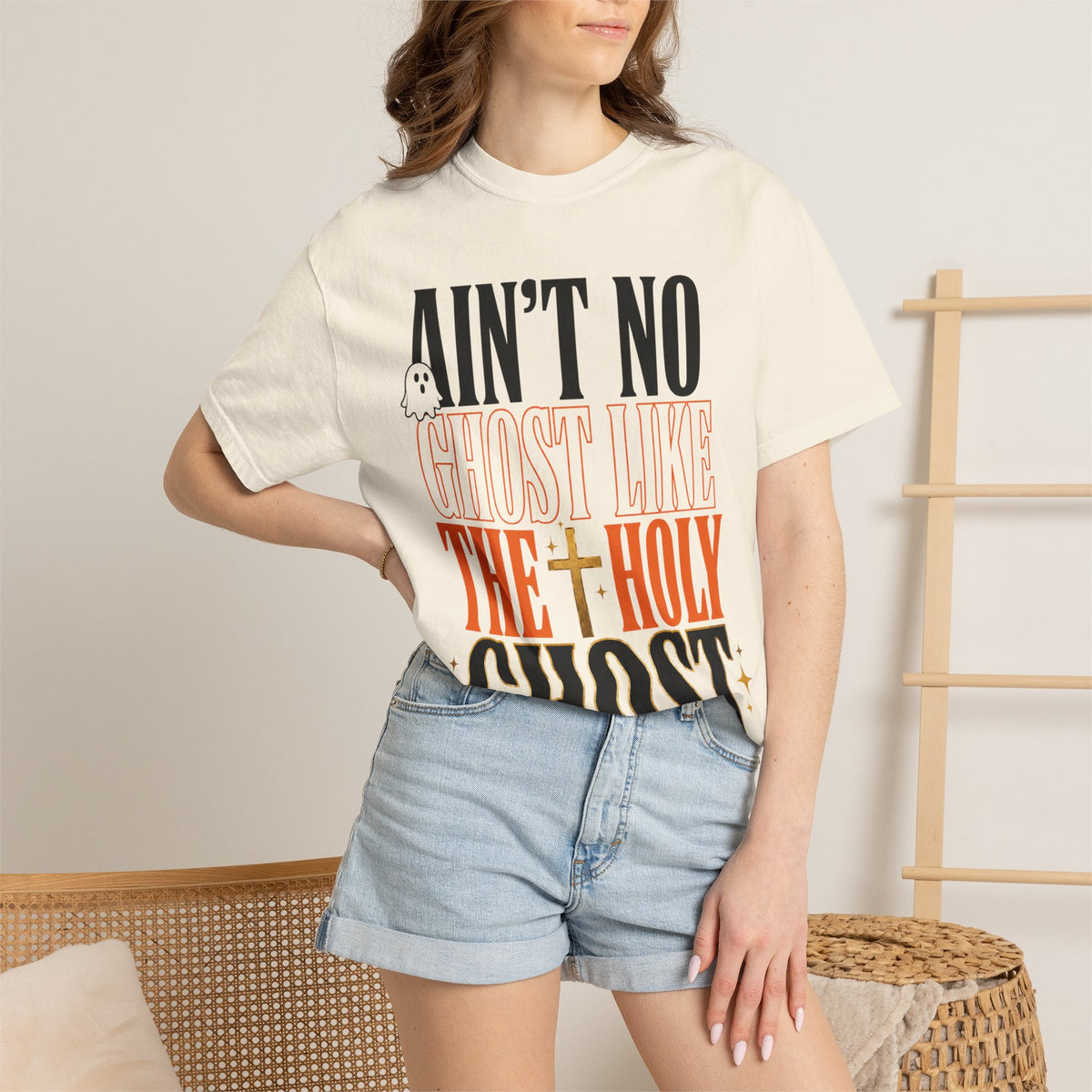 Woman wearing a cream graphic T shirt with bold text reading Ain't No Ghost Like the Holy Ghost paired with light denim shorts in a casual fashion setting