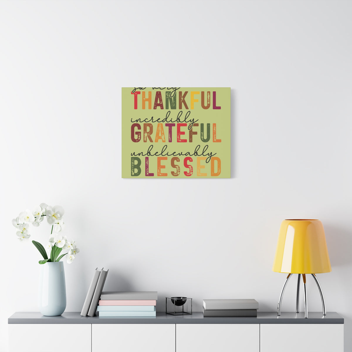 Distressed autumn wall art canvas with Thankful Grateful and Blessed lettering displayed above a modern console with books white orchid and yellow lamp