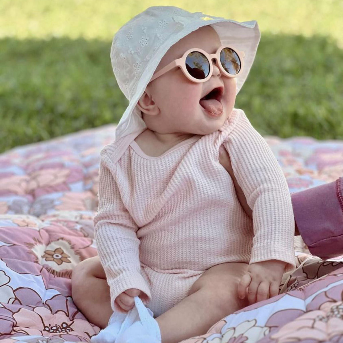 Baby in sunglasses and a sun hat sitting on a colorful floral quilted picnic blanket outdoors on grass