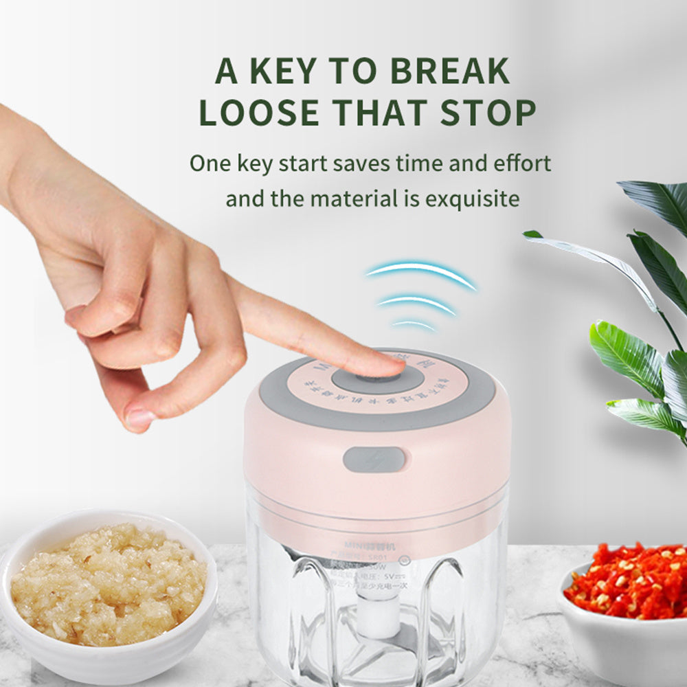 Compact electric garlic chopper with a hand pressing the top button, minced garlic in a bowl, and chopped red peppers nearby on a kitchen counter