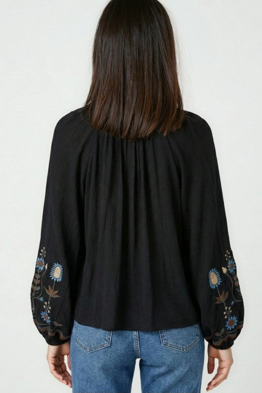 Back view of a woman wearing a loose black bohemian blouse with colorful floral embroidery on the sleeves and blue jeans from miss sparkling