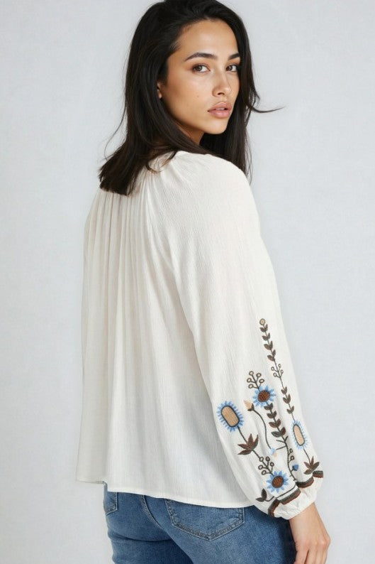 Woman wearing a loose ivory blouse with colorful embroidered floral details on the sleeve and blue jeans in a relaxed bohemian fashion look