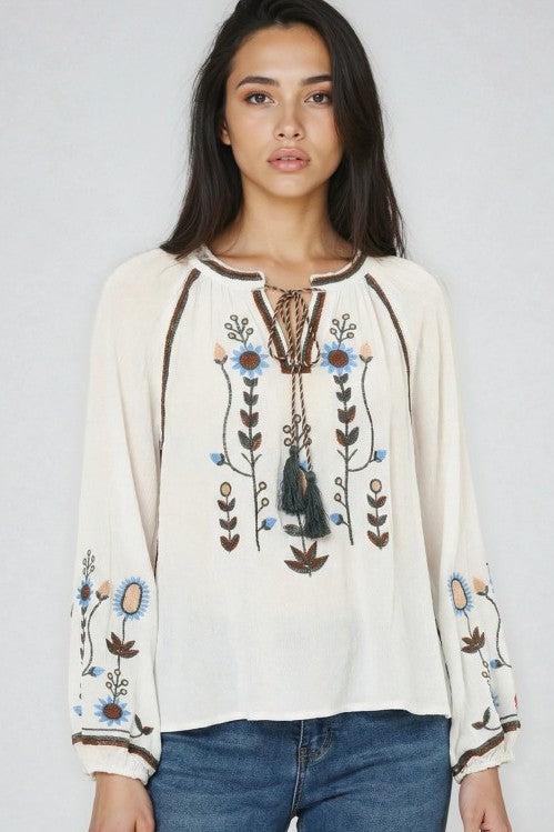 Woman wearing an ivory bohemian blouse with colorful embroidered flowers tassel tie neck and long sleeves paired with blue jeans against a light backdrop