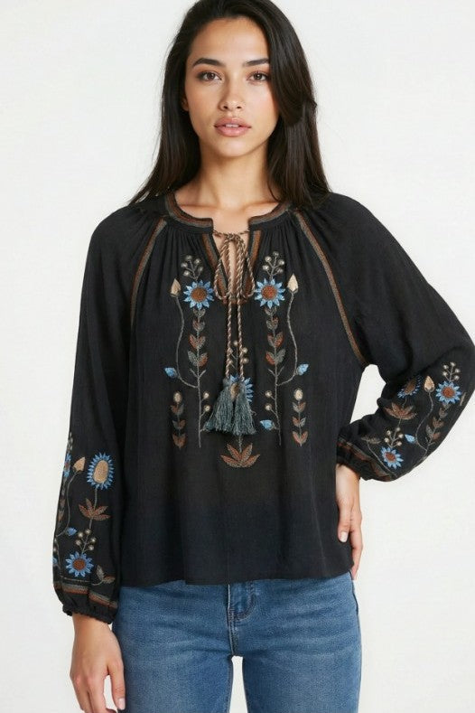 A woman models a black bohemian blouse with colorful floral embroidery long sleeves and a tassel tie neckline with blue jeans