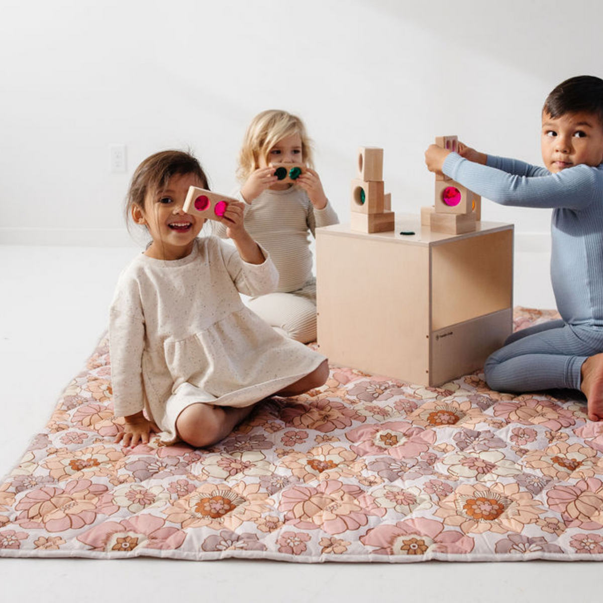 Three children play with wooden blocks and colorful toys on a pink floral quilted picnic blanket in a bright room from DC Toki Mats Home and beyond