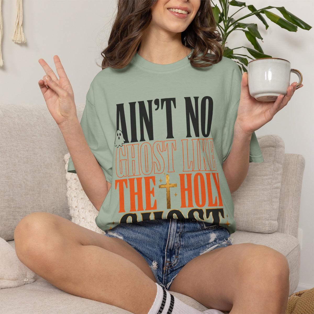 Woman wearing a sage green faith inspired graphic tee with bold black and orange lettering while seated on a sofa and holding a mug in a cozy home setting