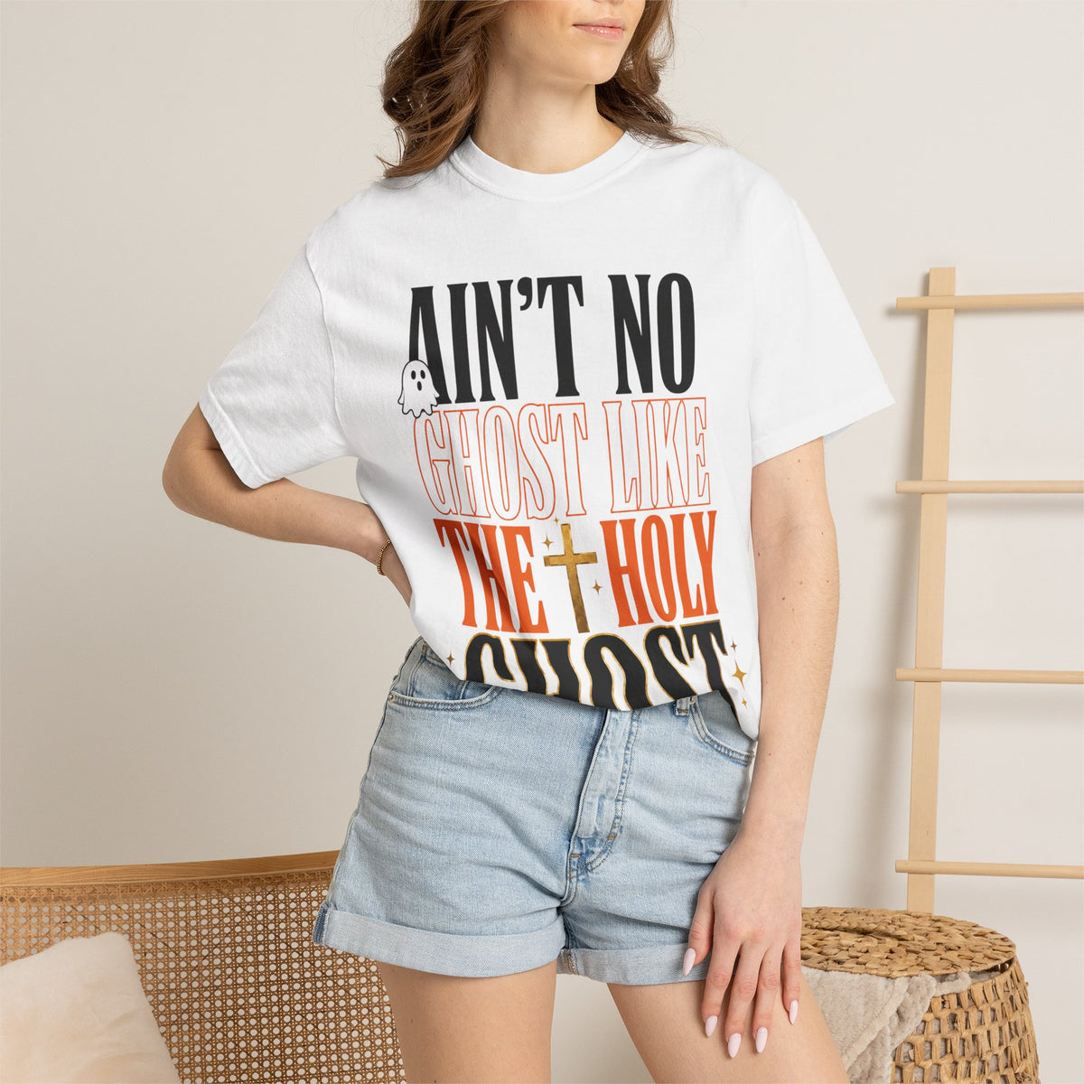 Woman wearing a white graphic T shirt featuring an Ain’t No Ghost Like the Holy Ghost design styled with denim shorts in a relaxed home setting