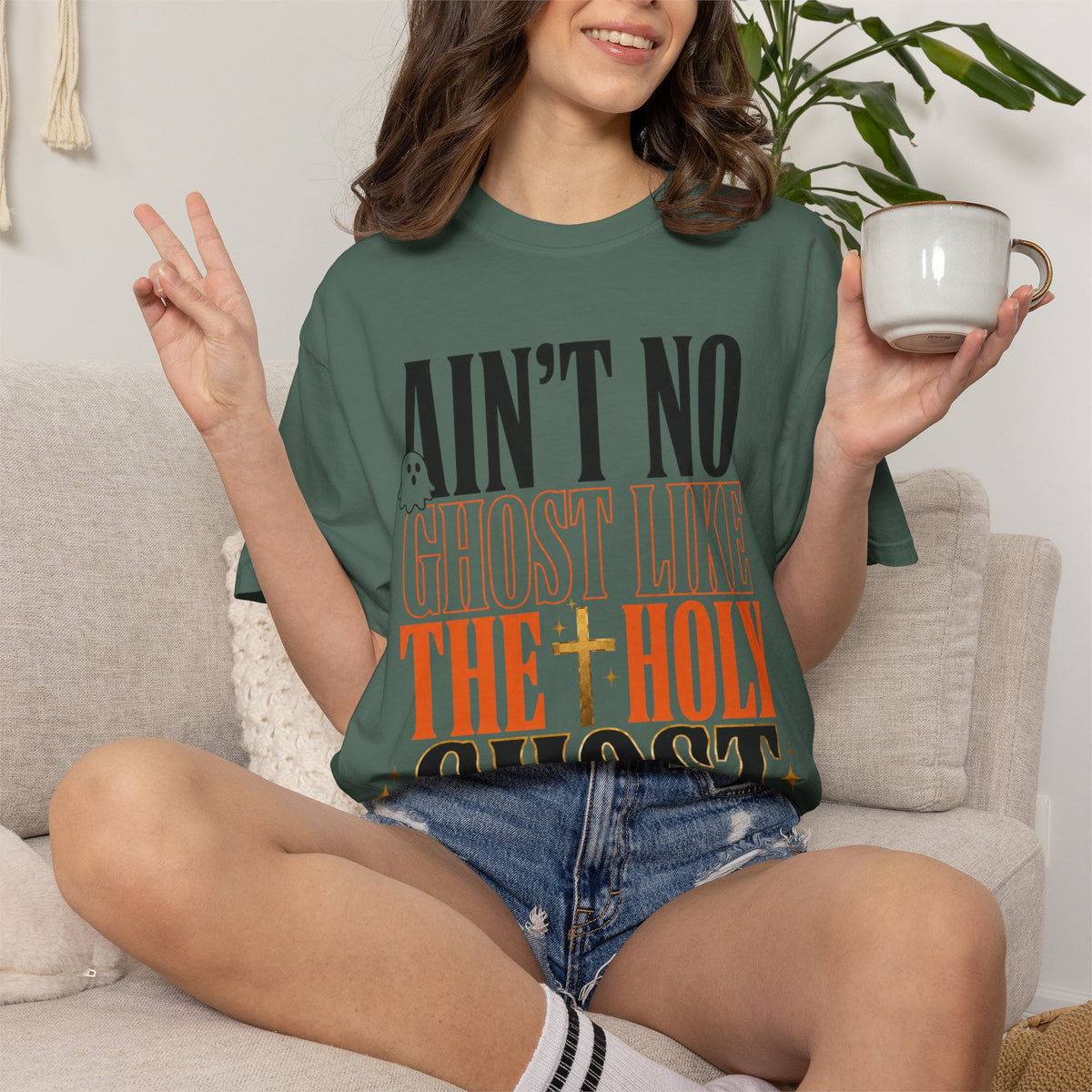 Woman wearing a forest green faith graphic T shirt with bold orange and black lettering while holding a mug and flashing a peace sign on a sofa