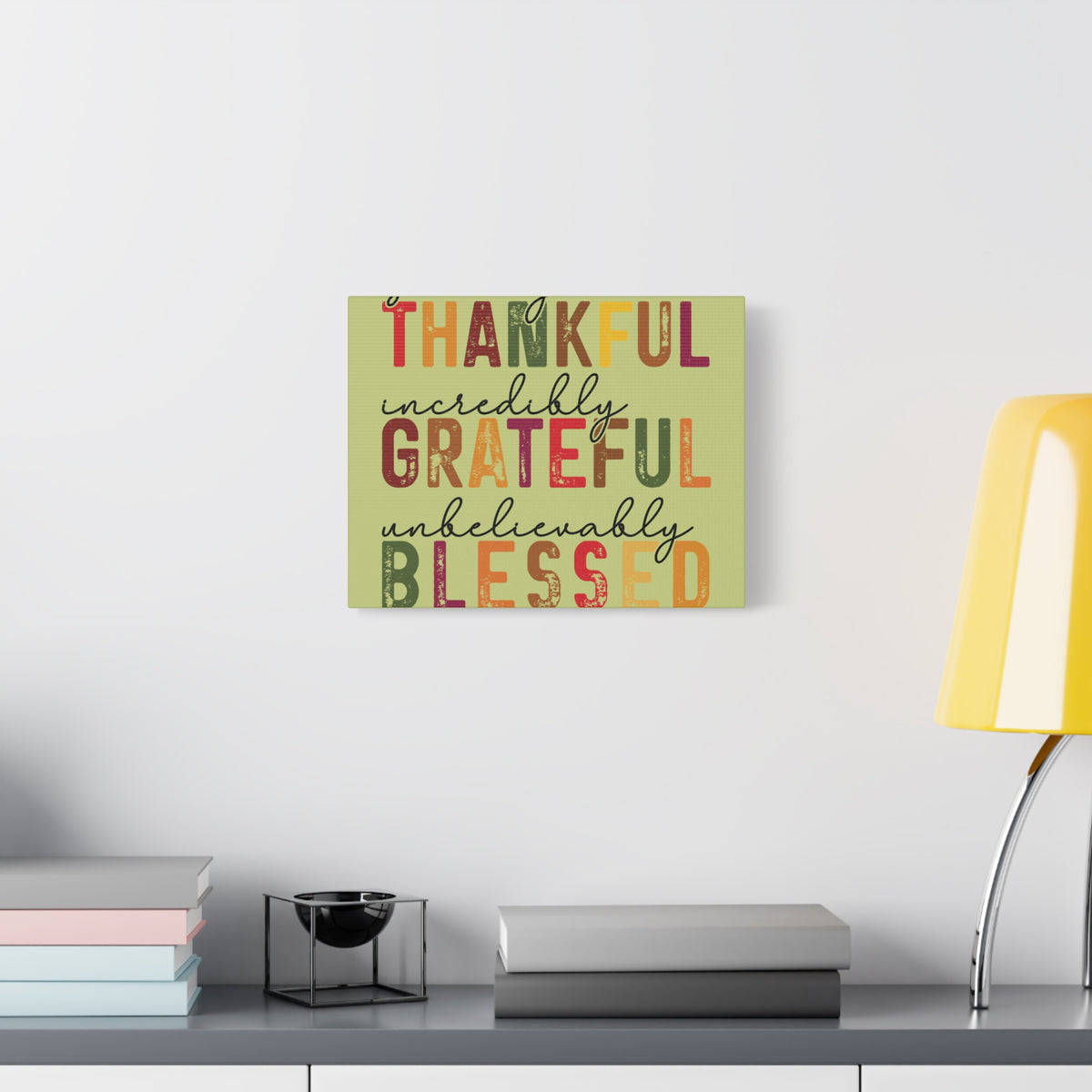 Autumn inspired matte canvas wall art featuring the words thankful grateful and blessed above a shelf with books and a yellow lamp from Printify Home and beyond