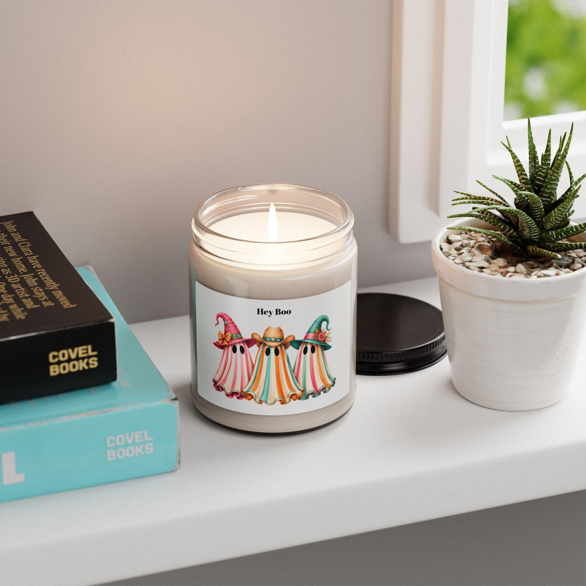 Lit scented soy candle in a glass jar featuring three colorful ghosts wearing cowboy hats on a windowsill beside books and a small succulent plant