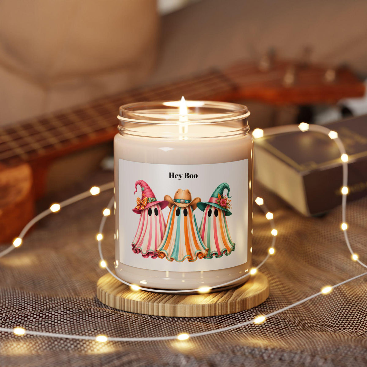 Lit glass candle with three colorful ghost illustrations wearing hats on the label surrounded by warm string lights and a guitar in the background