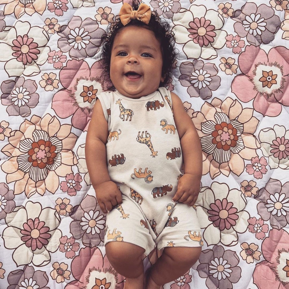 Smiling baby lying on a pink and mauve floral quilted blanket wearing an animal print romper and bow headband