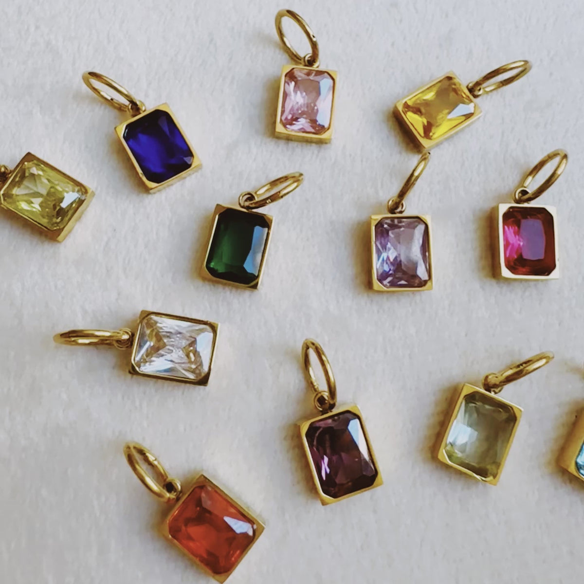 Assorted gold tone rectangular birthstone pendants with colorful gemstones displayed on white fabric from Ellison and Young for women's jewelry