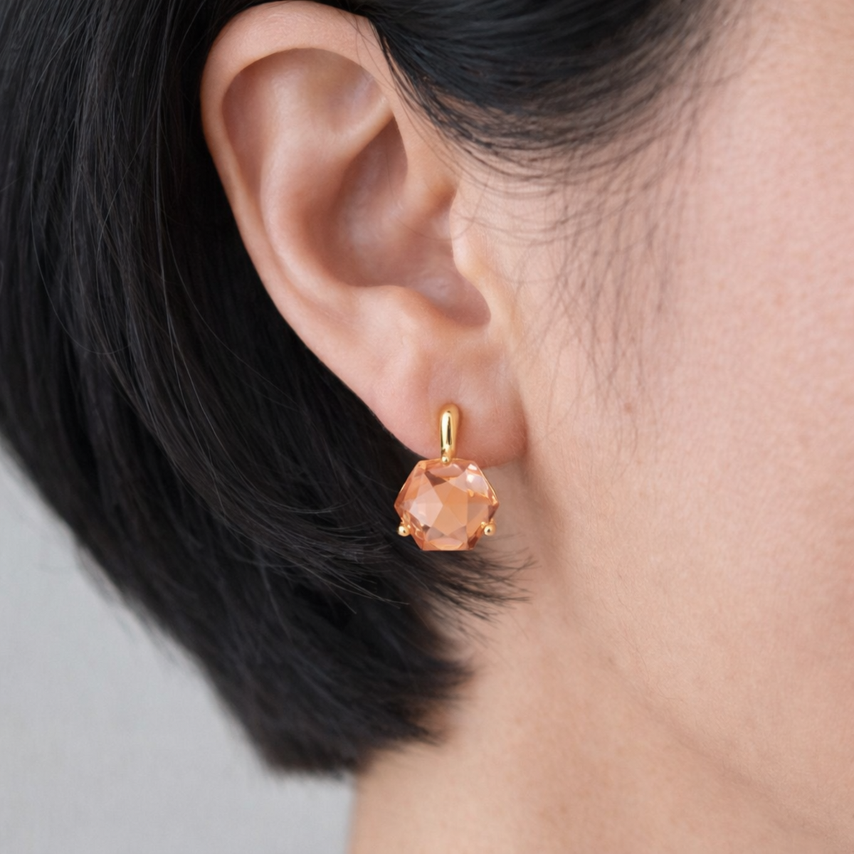 Close up of a faceted peach stone drop earring in a sculpted gold setting worn on the ear from Ellison + Young Jewelry for women's fashion
