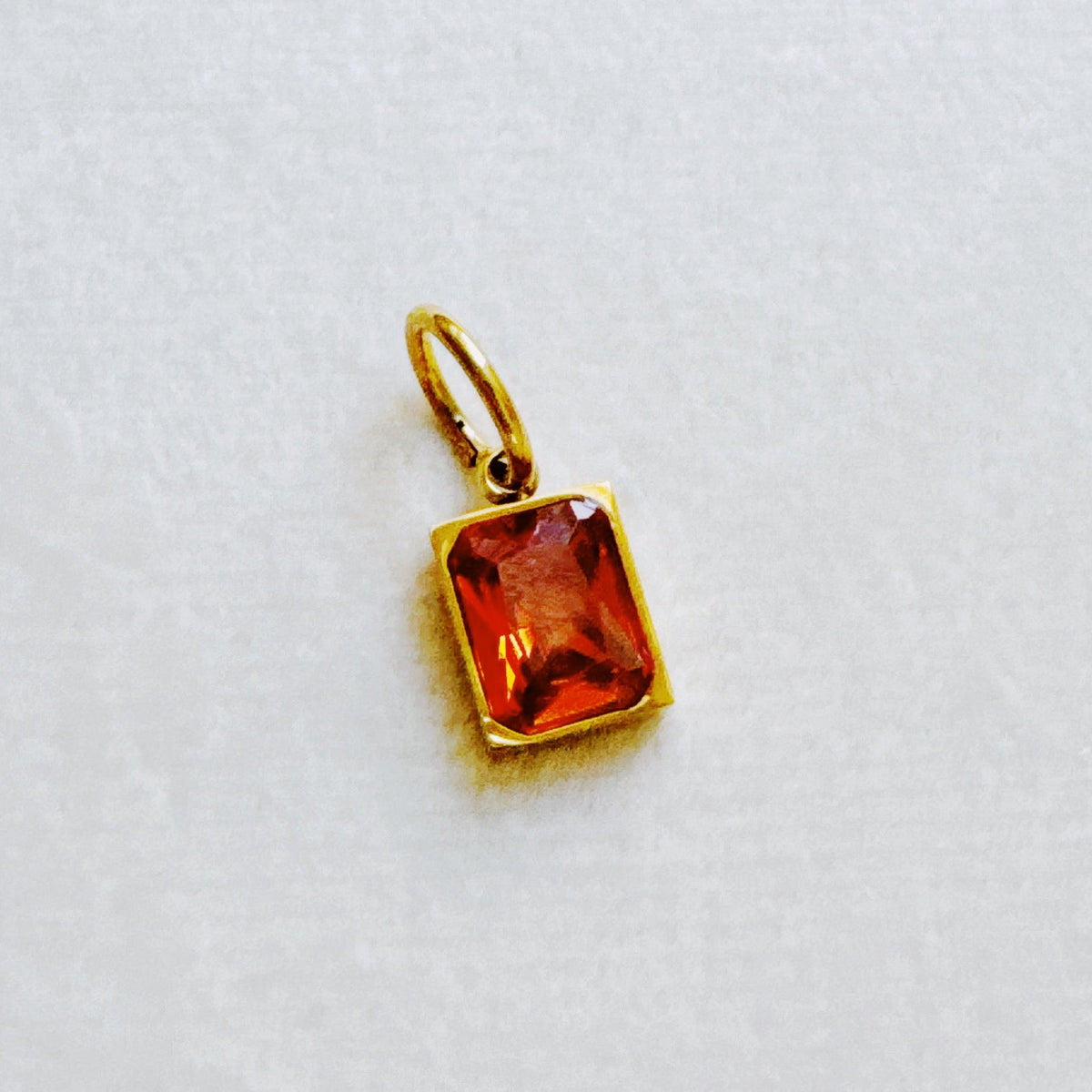 Gold tone birthstone charm with a faceted rectangular red orange gemstone on a white background for stylish everyday jewelry
