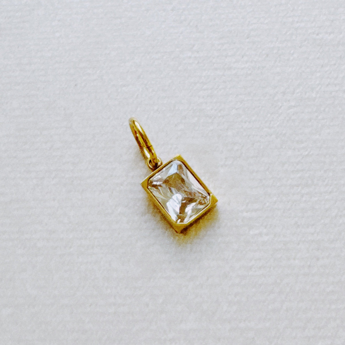 Gold rectangular birthstone pendant with a clear faceted charm displayed on white fabric from Ellison + Young jewelry collection