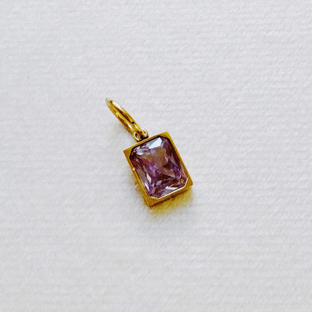 Gold tone rectangular purple birthstone pendant with faceted gemstone displayed on white fabric from Ellison and Young Best Sellers Jewelry collection