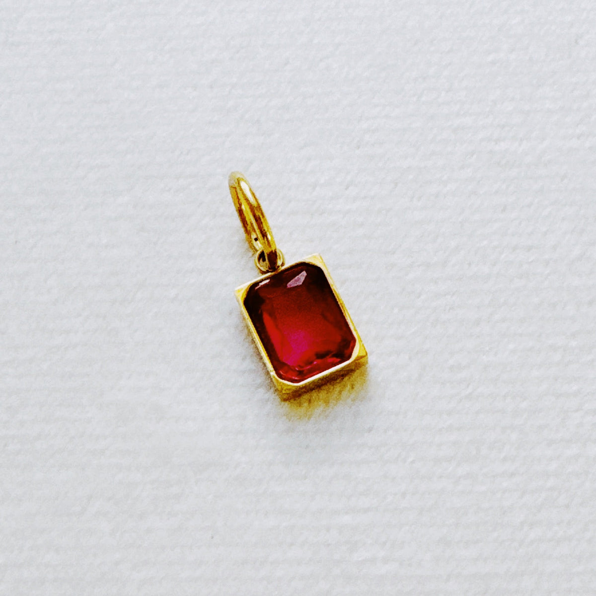 Red rectangular birthstone pendant in a gold tone setting on white fabric by Ellison + Young from the Best Sellers Jewelry collection