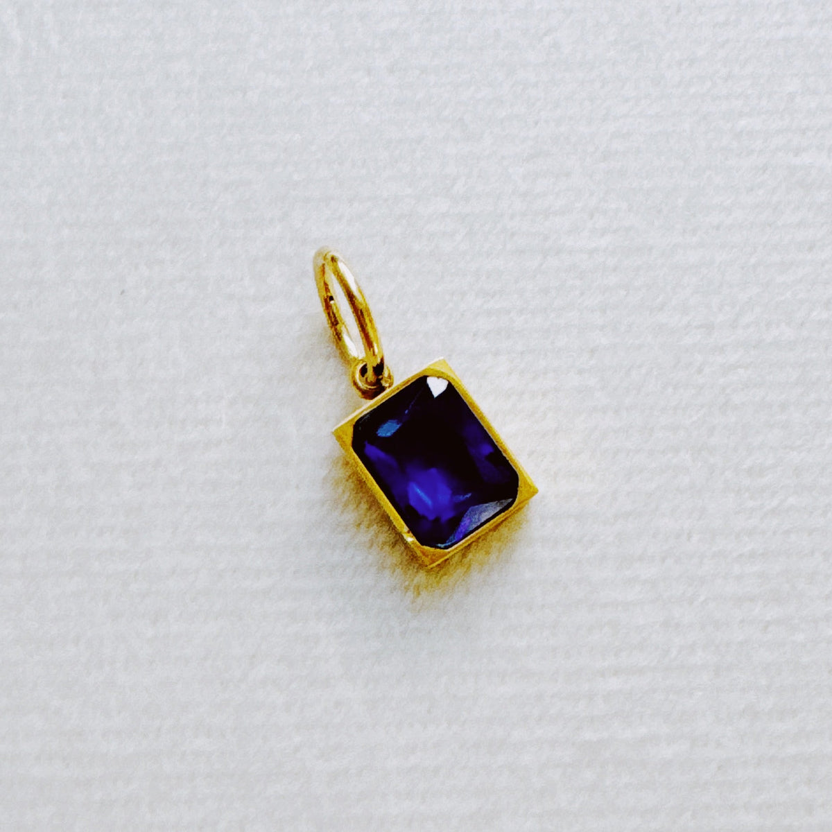 Gold rectangular pendant with a deep blue faceted birthstone charm displayed on a white textured background from Ellison + Young