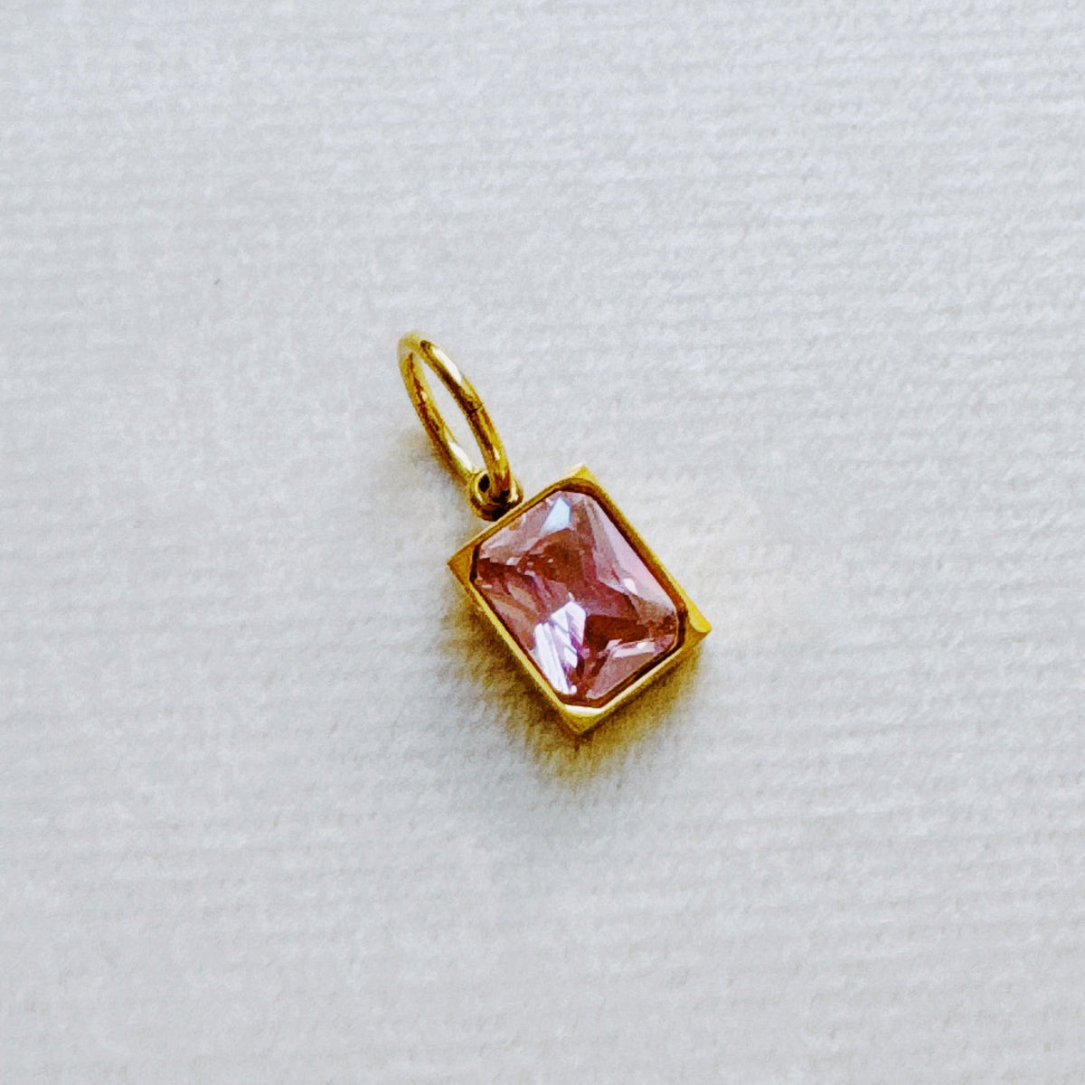 Gold tone rectangular pendant with a faceted pink birthstone charm displayed on soft white textured fabric for a delicate women's jewelry accessory