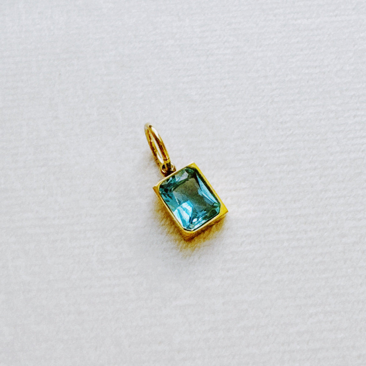 Gold tone rectangular blue green birthstone pendant with a polished loop on white textured fabric from Ellison and Young Best Sellers Jewelry collection