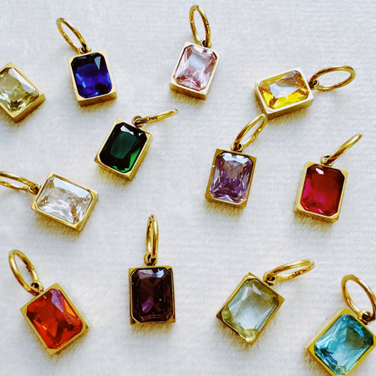 Assorted colorful rectangular birthstone pendants in gold settings arranged on white fabric from Ellison + Young for Best Sellers and Jewelry