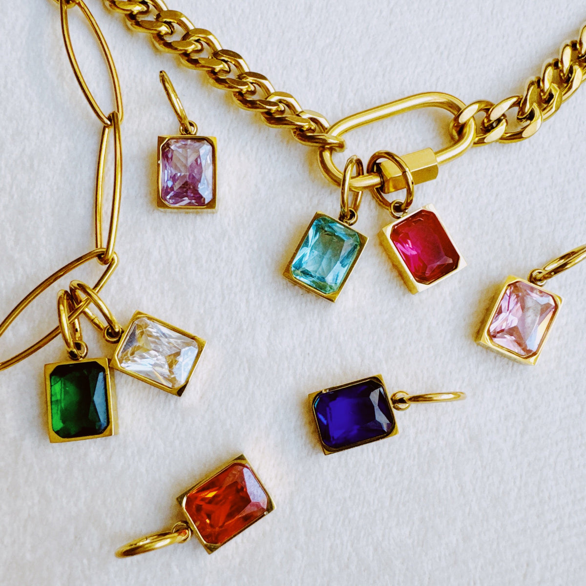 Gold tone pendants with colorful rectangular birthstone charms in pink blue red green purple and orange arranged on white fabric from the Jewelry collection