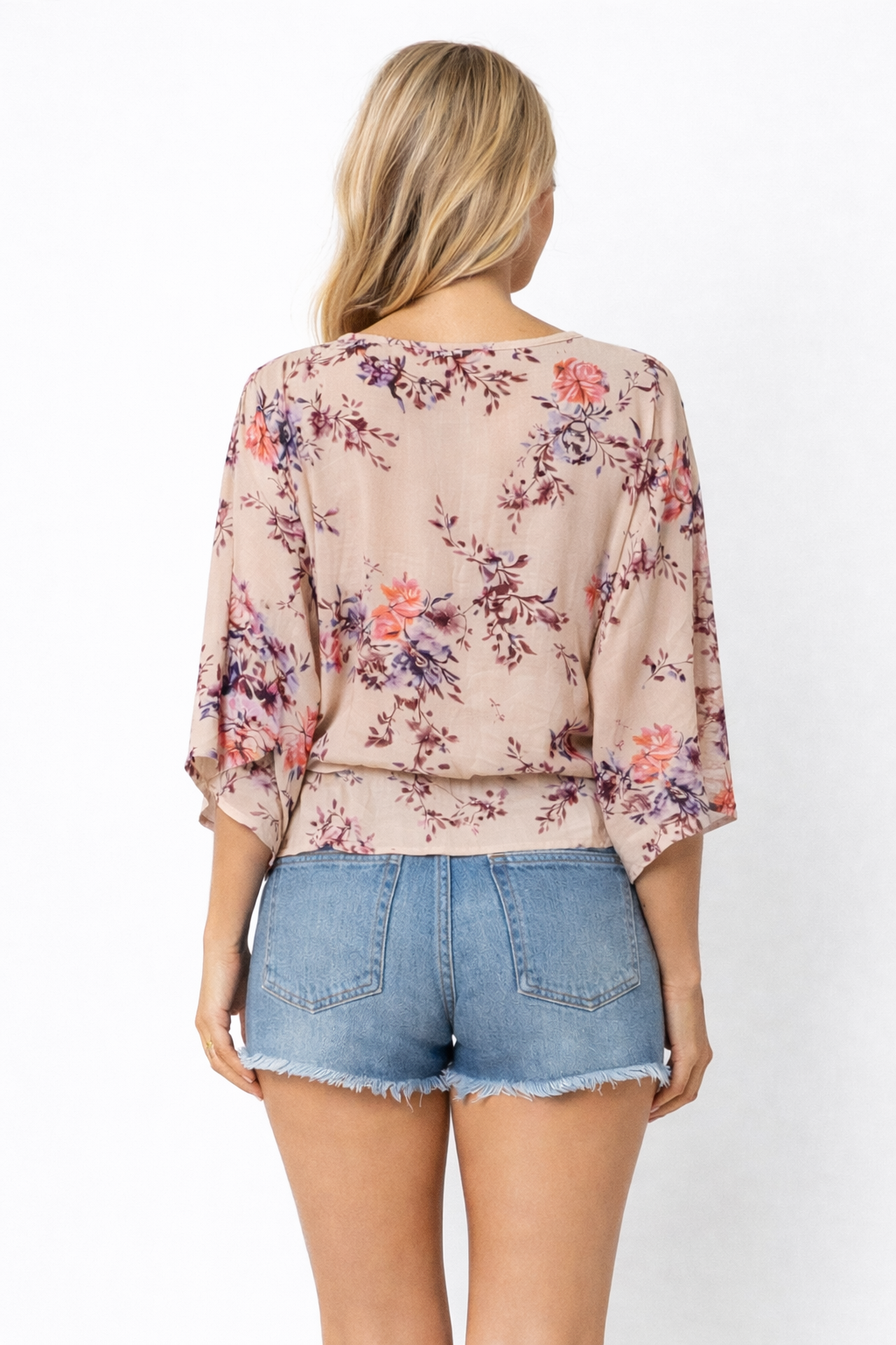 Back view of a woman wearing a soft beige floral wrap blouse with three quarter sleeves and a drawstring hem paired with frayed denim shorts