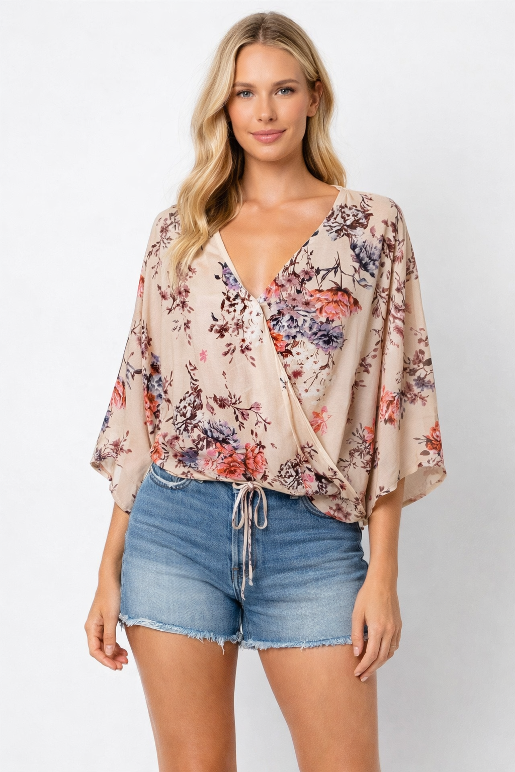 Woman wearing a beige floral wrap blouse with pink coral and mauve flowers and an adjustable drawstring hem styled with denim shorts
