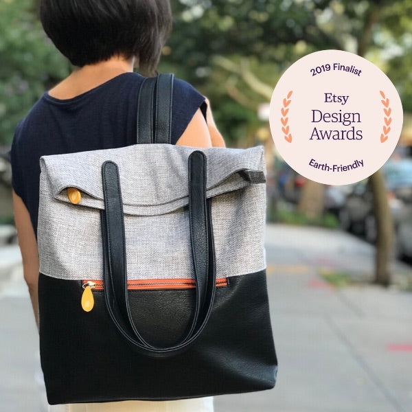 Gray and black convertible backpack tote worn outdoors with shoulder straps and orange zipper accents beside an Etsy Design Awards finalist badge