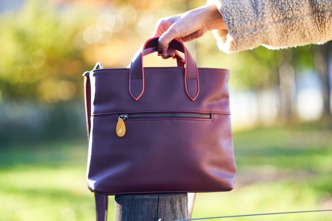 Women's burgundy satchel bag with top handles and a front zip pocket held outdoors by a hand from DC-Canopy Verde in Bags and Belts