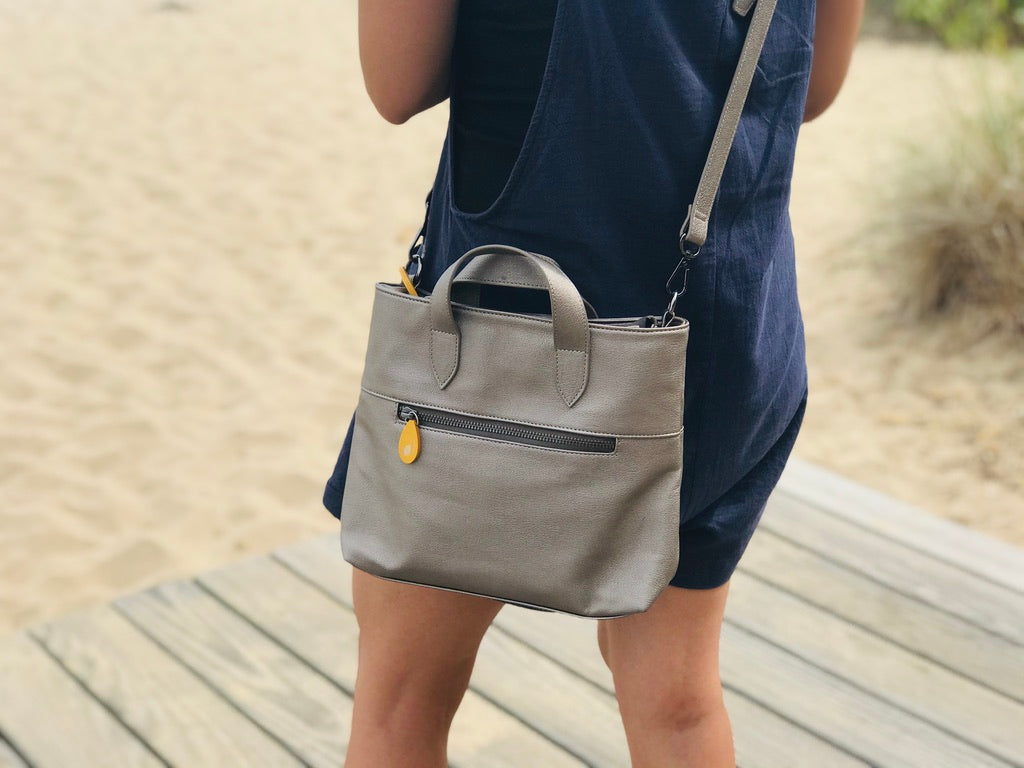 Woman wearing a gray crossbody satchel with top handles and front zip pocket on a boardwalk from DC Canopy Verde bags and belts collection