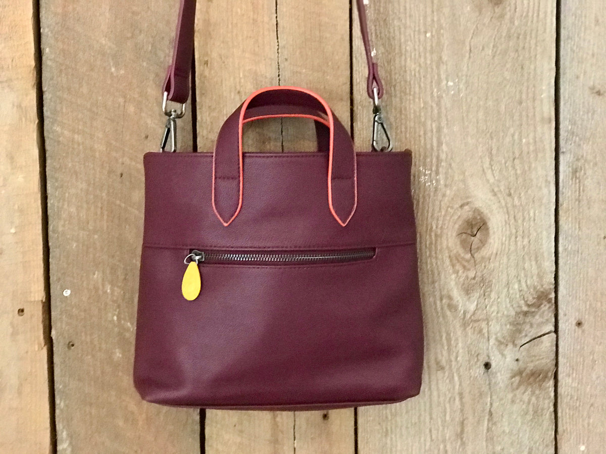 Burgundy crossbody bag with top handles and front zip pocket displayed against rustic wood for stylish women's bags and accessories