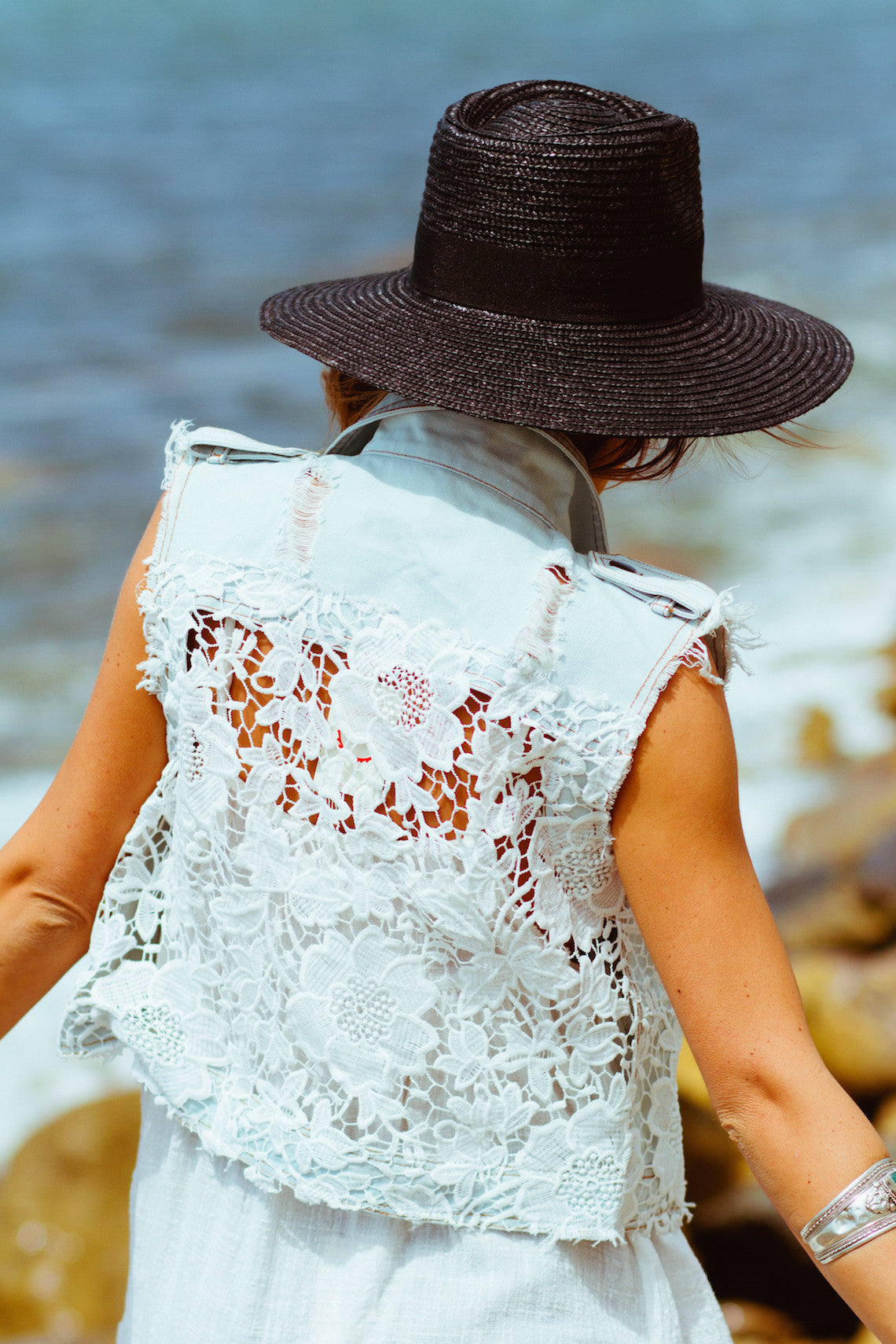 Woman wearing a white lace back vest with frayed edges and a wide brim black hat outdoors near the water in stylish women's fashion attire
