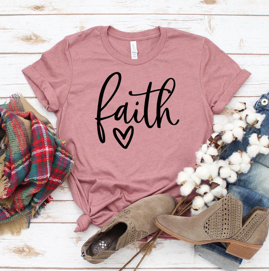 Dusty rose women's graphic T shirt featuring black Faith lettering and a heart design styled with plaid scarf and tan ankle boots on a rustic wood background