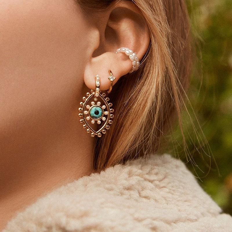 Close up of a woman wearing an ornate gold toned drop earring with pearl accents and a turquoise center plus a beaded ear cuff from Drizzle and Tizzle Jewelry