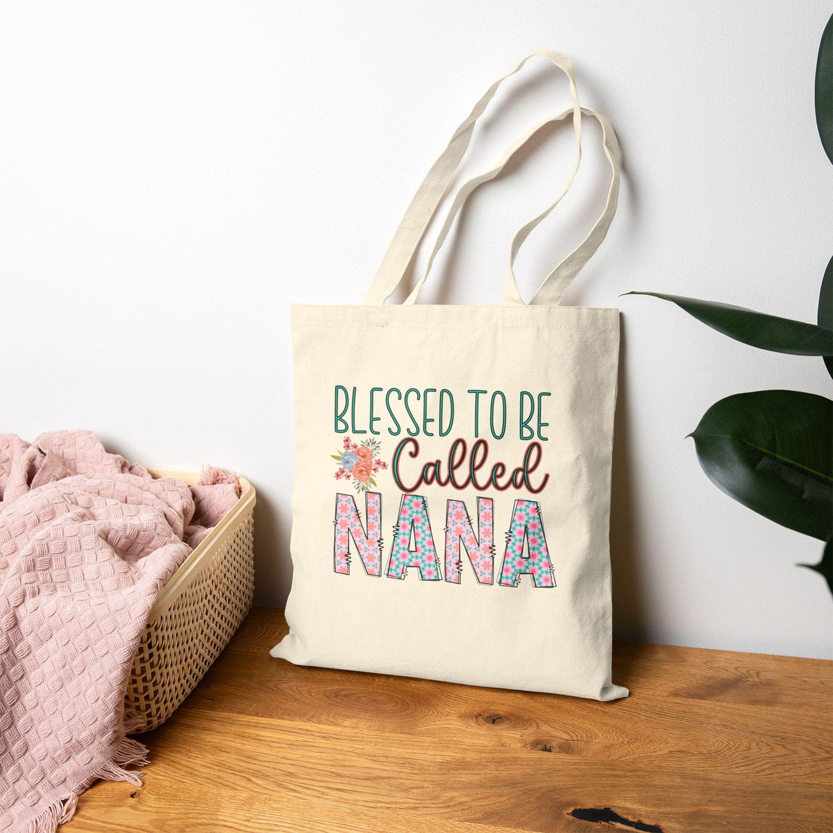 Cotton canvas tote bag with colorful Blessed to Be Called Nana lettering beside a woven basket and pink blanket on a wooden table