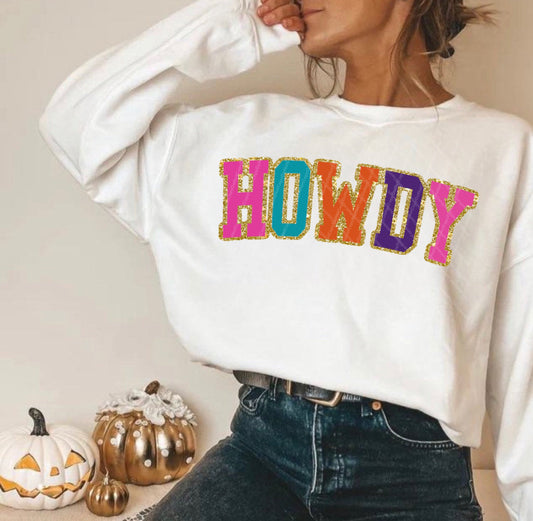 Woman wearing a white Howdy glitter print crewneck sweatshirt with colorful varsity lettering and jeans in a cozy pumpkin decorated setting