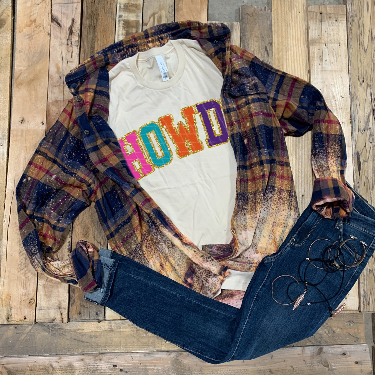 Women's casual outfit featuring a cream HOWDY graphic tee under a plaid cardigan with dark jeans and eyeglasses on a rustic wood floor