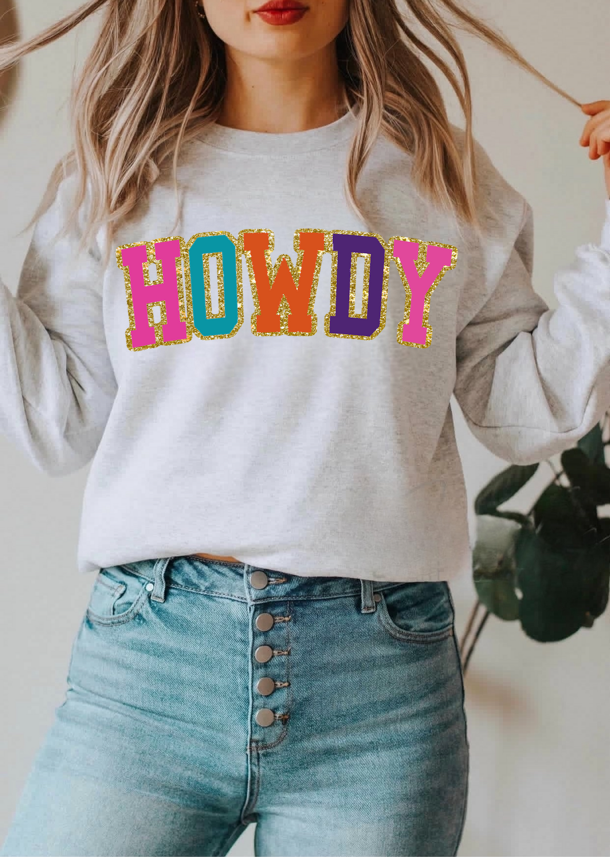 Woman wearing a light gray crewneck sweatshirt with a colorful glitter HOWDY graphic and high waisted jeans from DC-Blonde Tees women's fashion graphic apparel