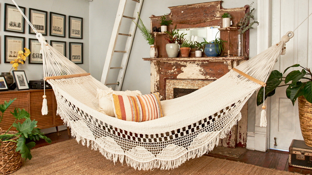 Cream cotton hammock with fringed macrame detailing and striped pillows in a cozy living room home decor setting