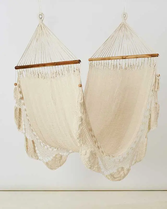 Two cream cotton hammocks with wooden spreader bars and delicate knotted fringe hanging against a light background for relaxed home decor