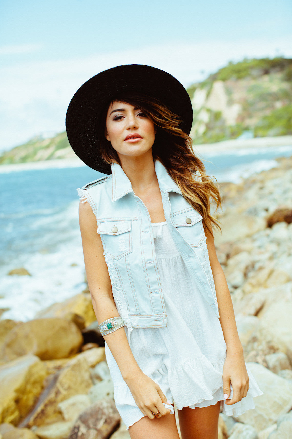 Woman wearing a light denim vest over a white outfit with a black wide brim hat beside a rocky seaside shore